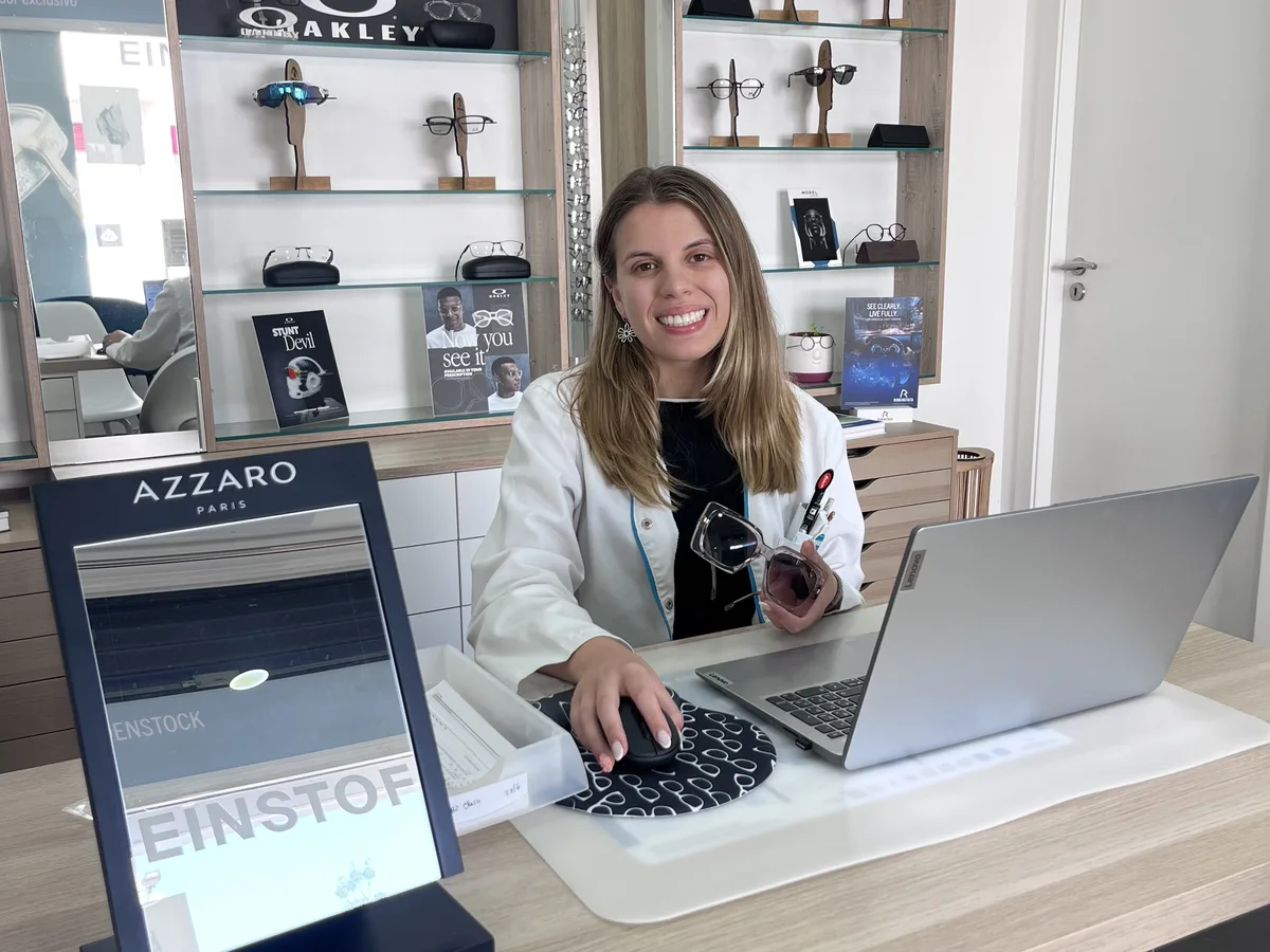 Bruna Ferreira em atendimento na mesa de trabalho do Centro Óptico da Torre, com expositores de óculos de sol ao fundo